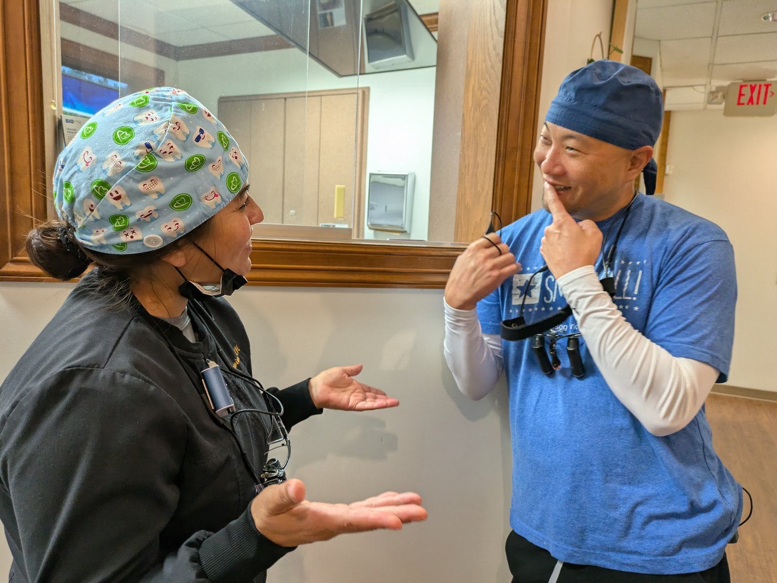 A dentist in a blue shirt and an assistant in a tooth-print cap laughing in the hallway