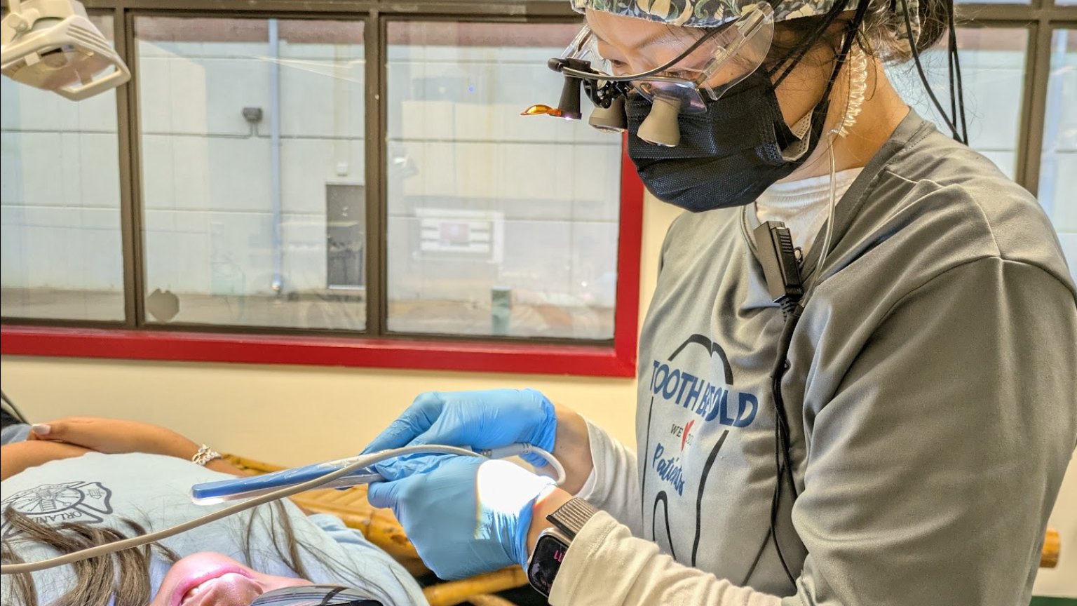 A hygienist in loupes and a floral cap working at the chair beside a reclined patient