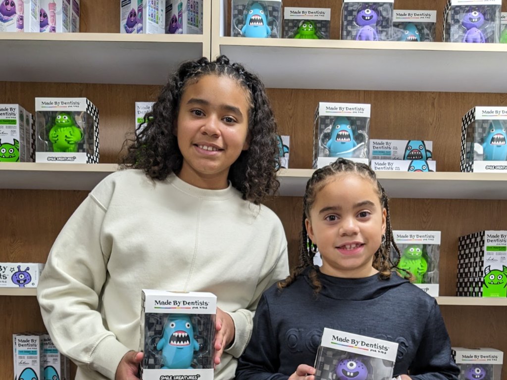 Two smiling girls holding toothbrush toys in front of a shelf of children's oral care products