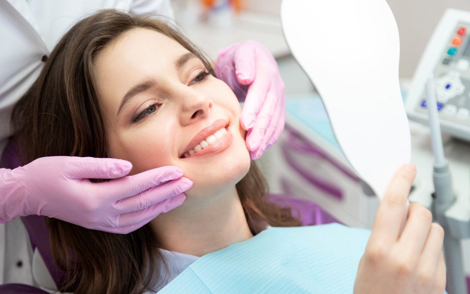 A patient in the chair smiling at her reflection in a hand mirror as the dentist holds her jaw