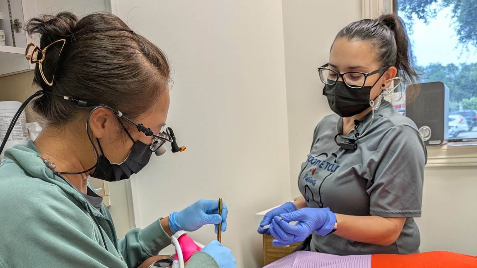 A hygienist in loupes working on a patient while an assistant hands her an instrument