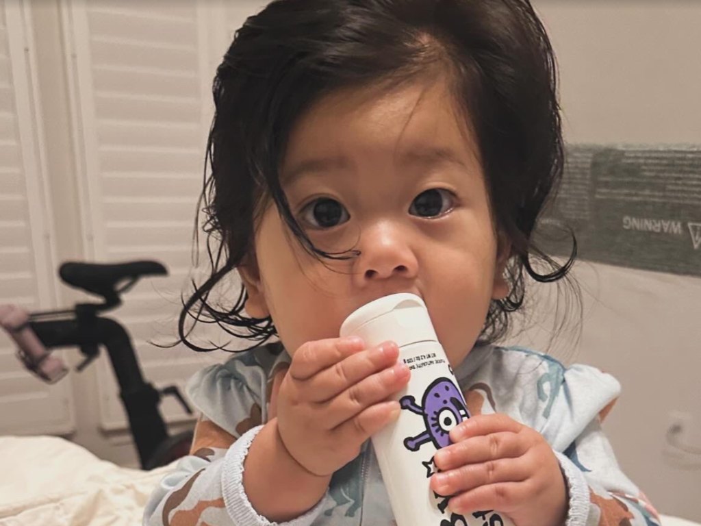 A baby in pyjamas sitting on a bed, chewing on the end of a toothpaste tube