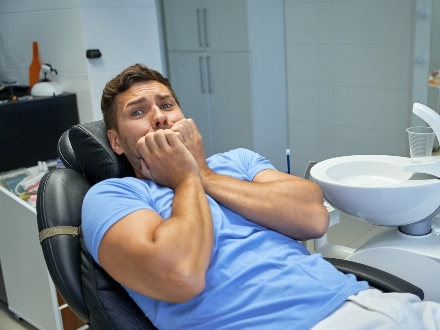 A man in a dental chair with his fists pressed to his mouth, eyes wide with fear