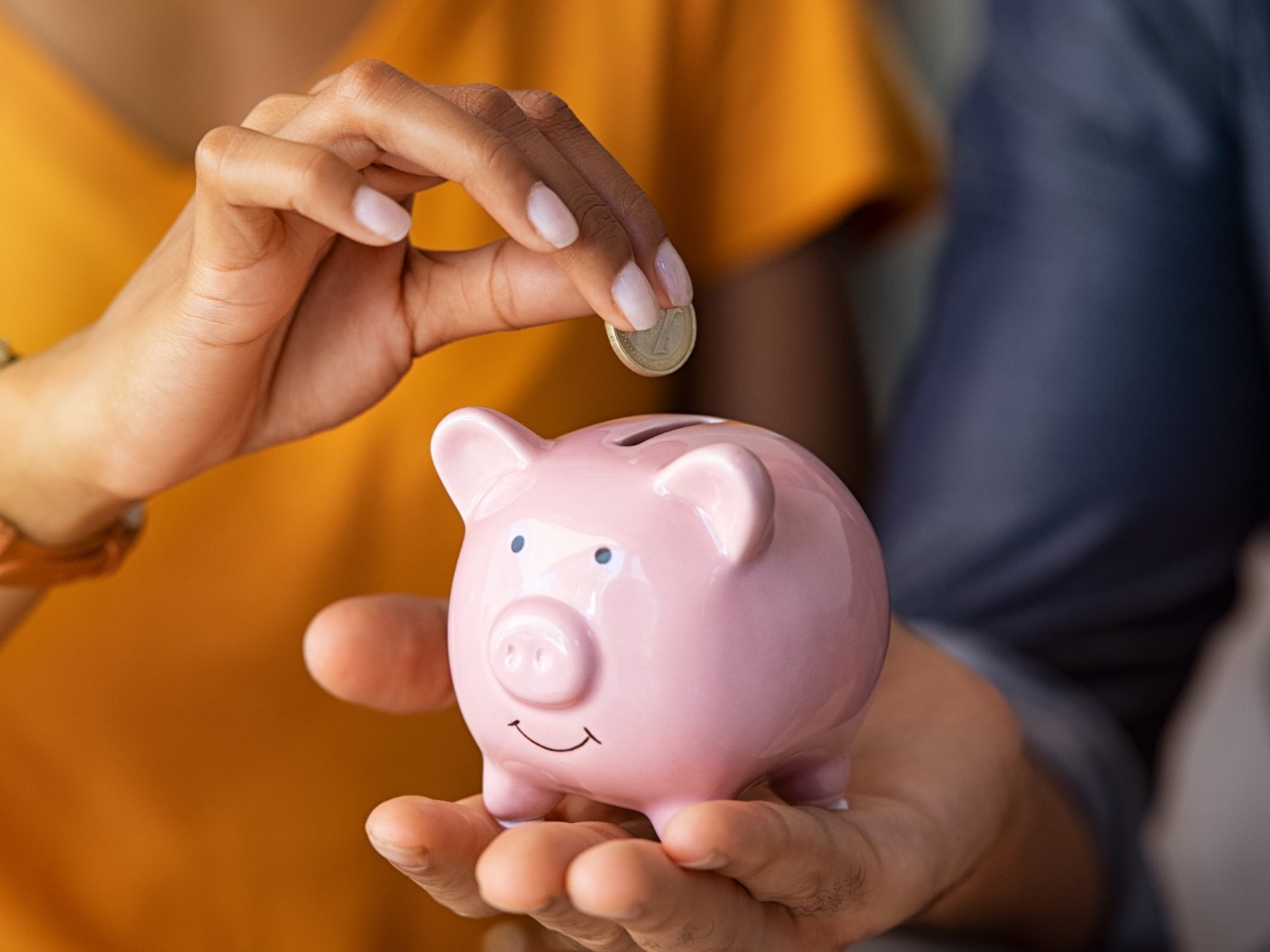 A coin being dropped into a pink piggy bank held in an open hand
