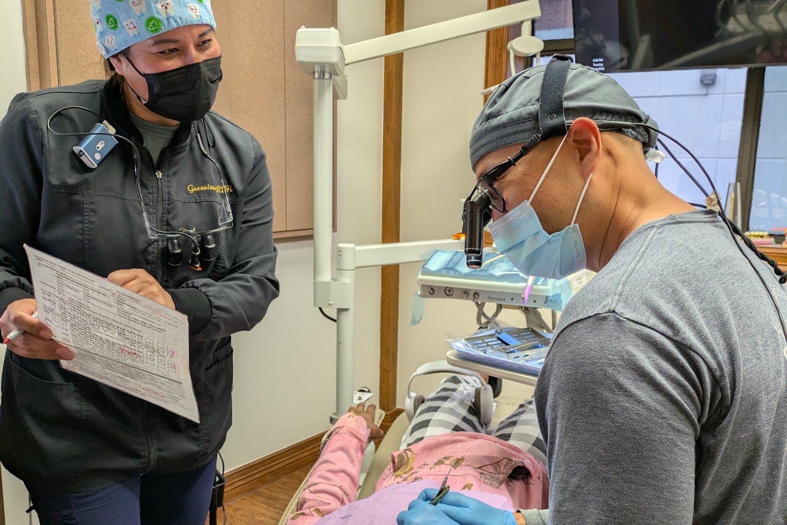 The dentist treating a patient while a hygienist holds the chart